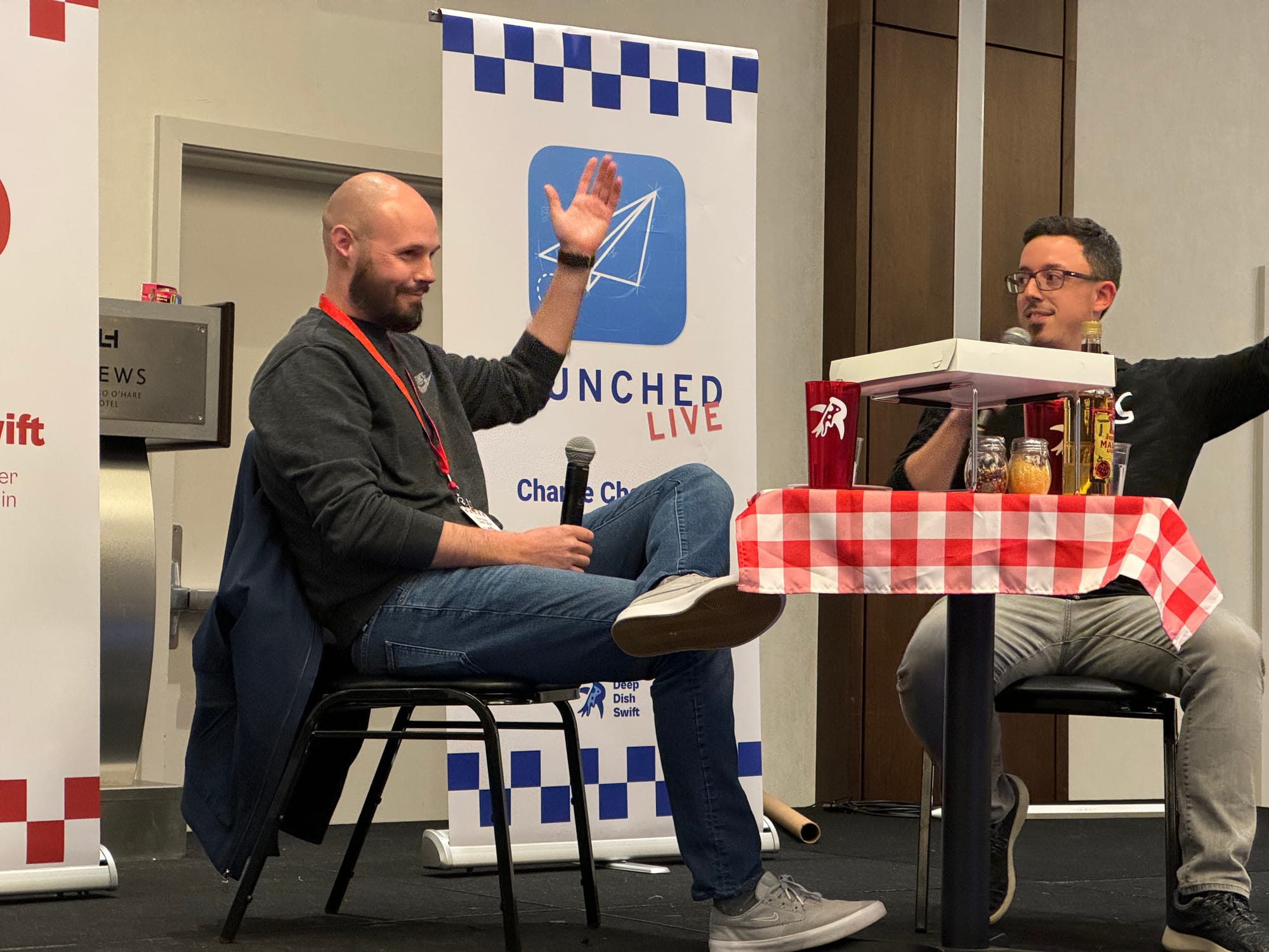 Sean and Charlie with mics in hand, sitting at a mock pizza table, podcasting on stage at Deep Dish Swift. There is a “Launched Live” banner in the background.